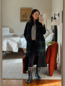 A girl wearing a black wool coat with a grey sweater and green plaid scarf with black Hunter rain boots